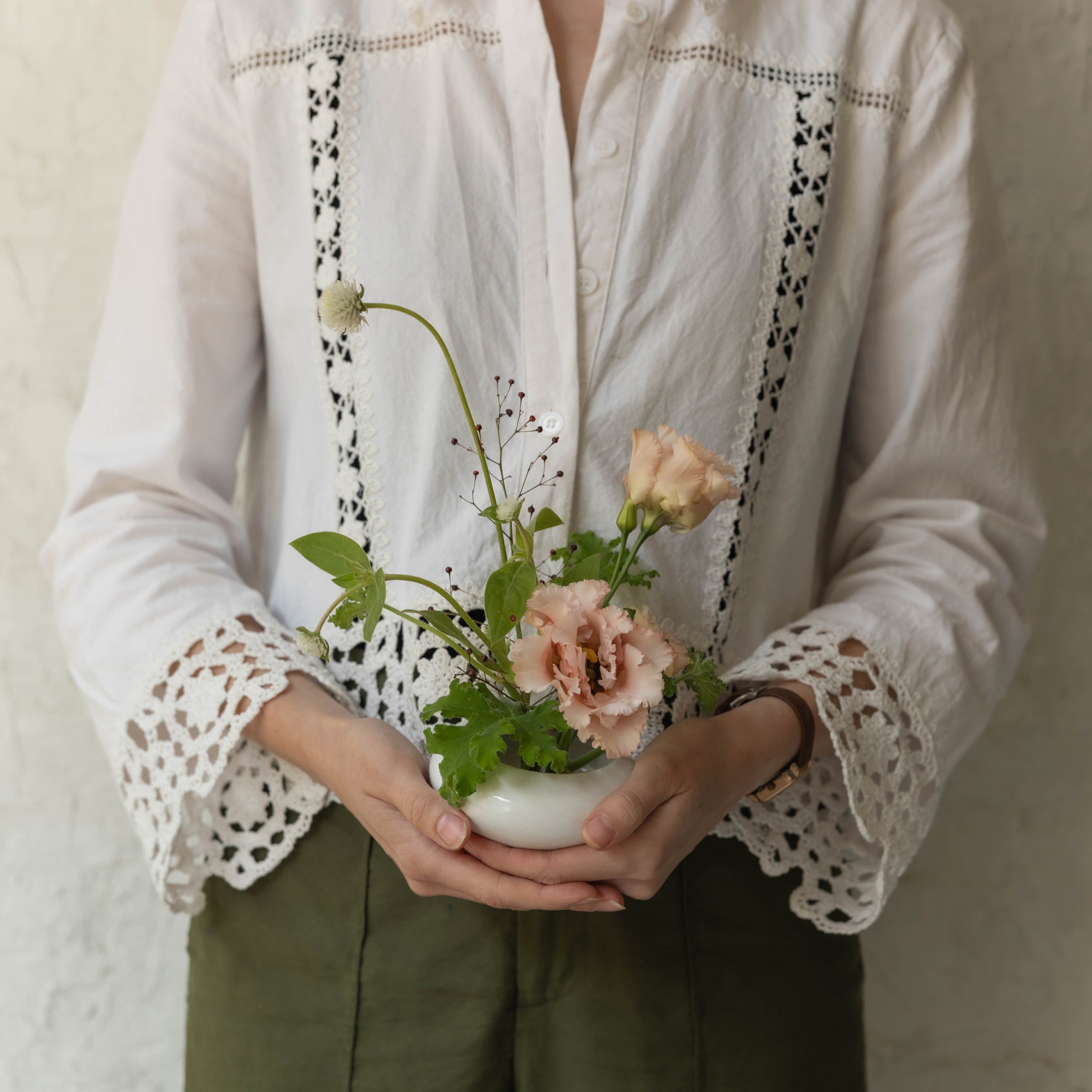 Person holding a small bouquet of flowers wearing a white blouse with lace details and green pants.