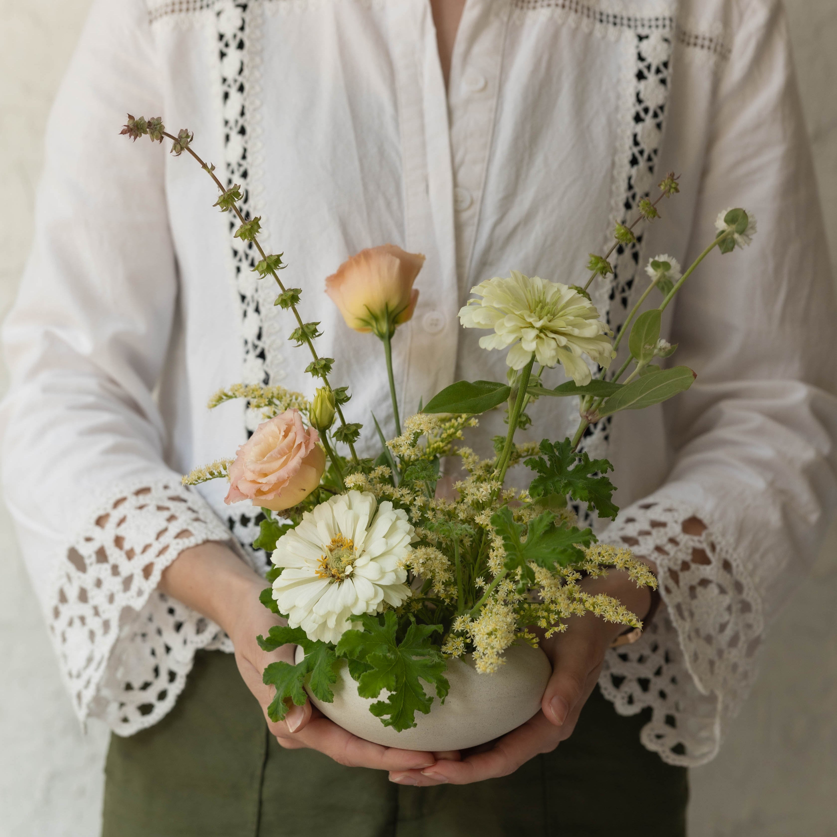 Person holding a small bouquet of flowers wearing a white blouse with lace details.