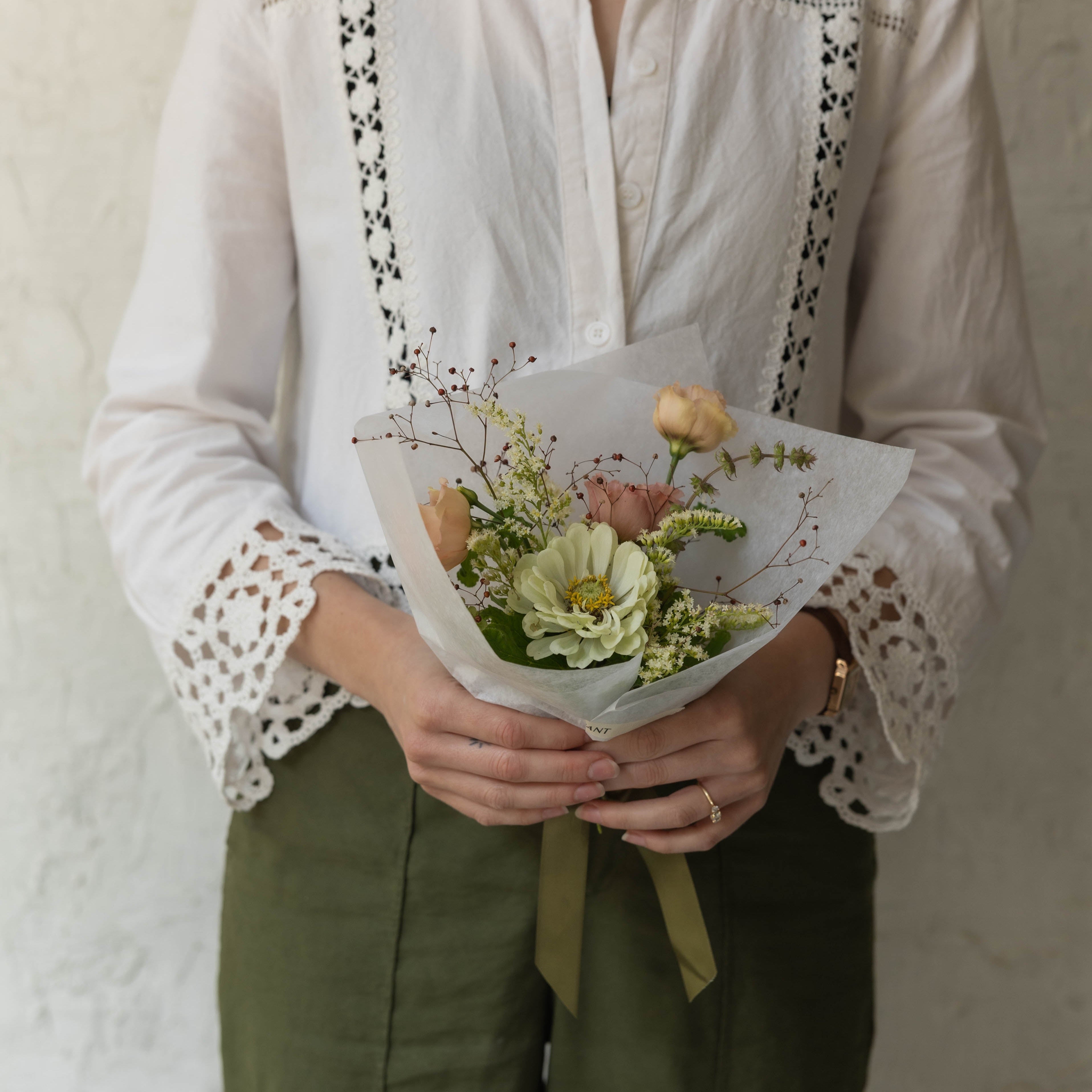 Person wearing a white blouse with lace details and holding a bouquet of flowers against a plain background