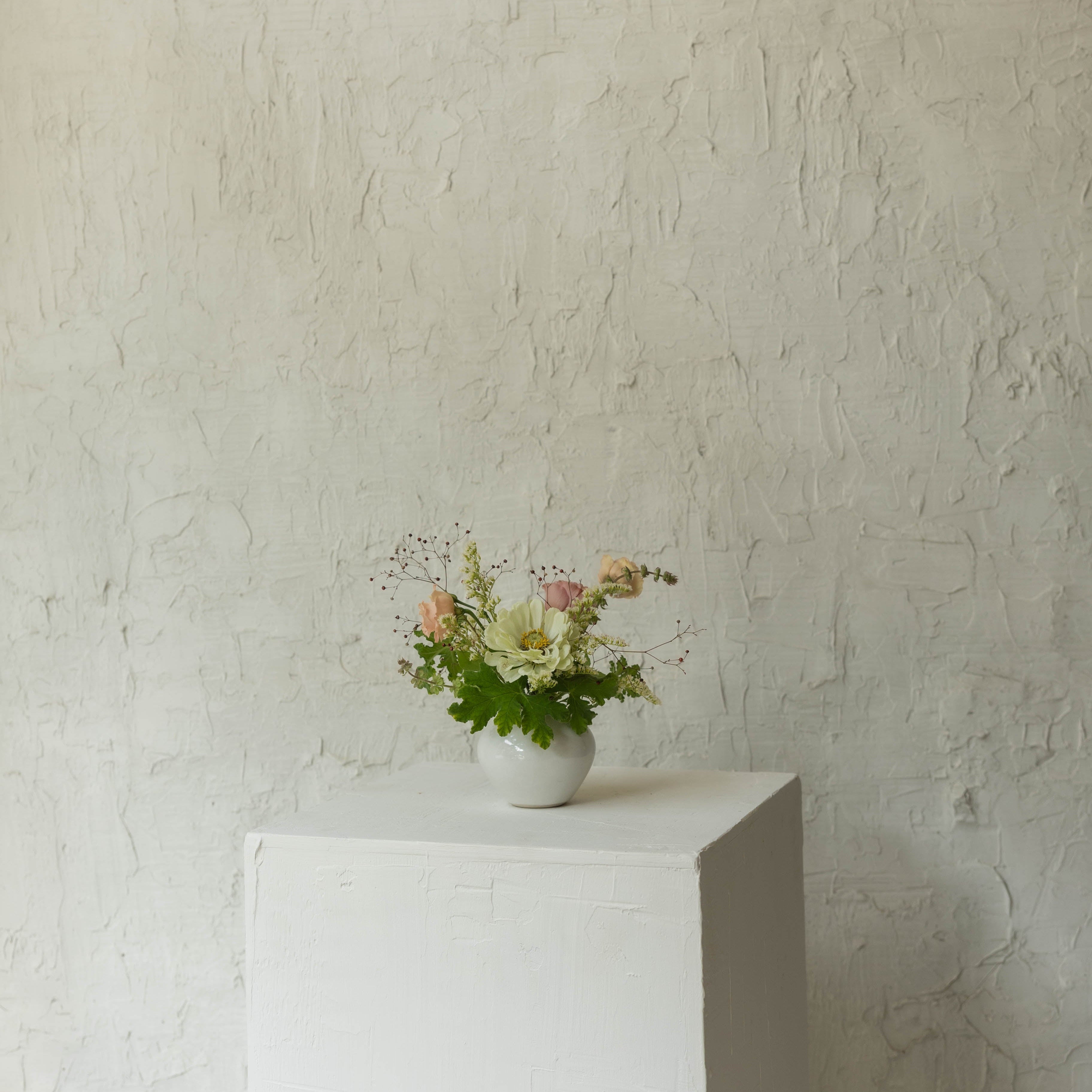 Small bouquet of flowers in a white vase on a white pedestal against a textured beige wall.