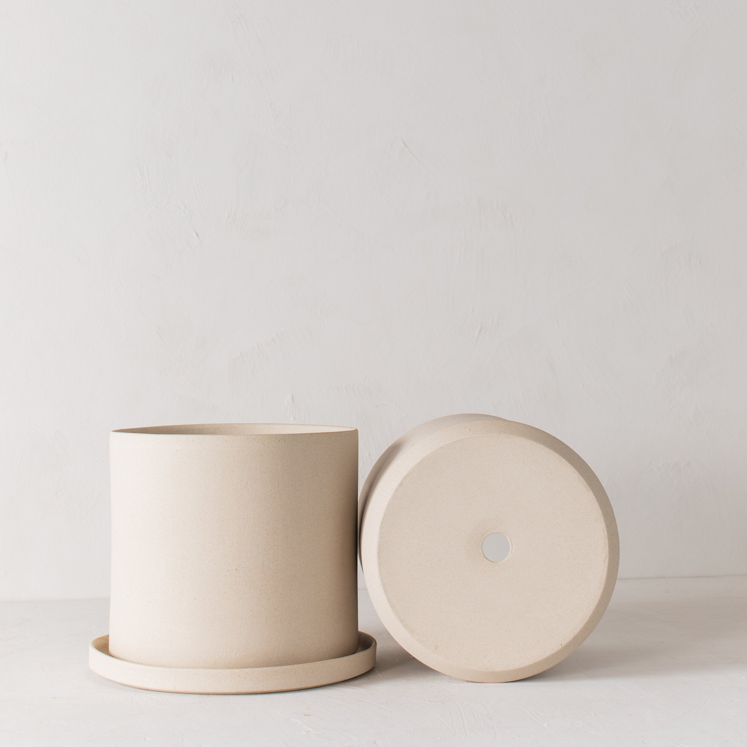 Two stoneware 10 inch ceramic planters with bottom drainage dish, one on its side with a drainage hole. Staged on a white plaster textured tabletop against a plaster textured white wall. Designed and sold by Convivial Production, Kansas City Ceramics.  Edit alt text