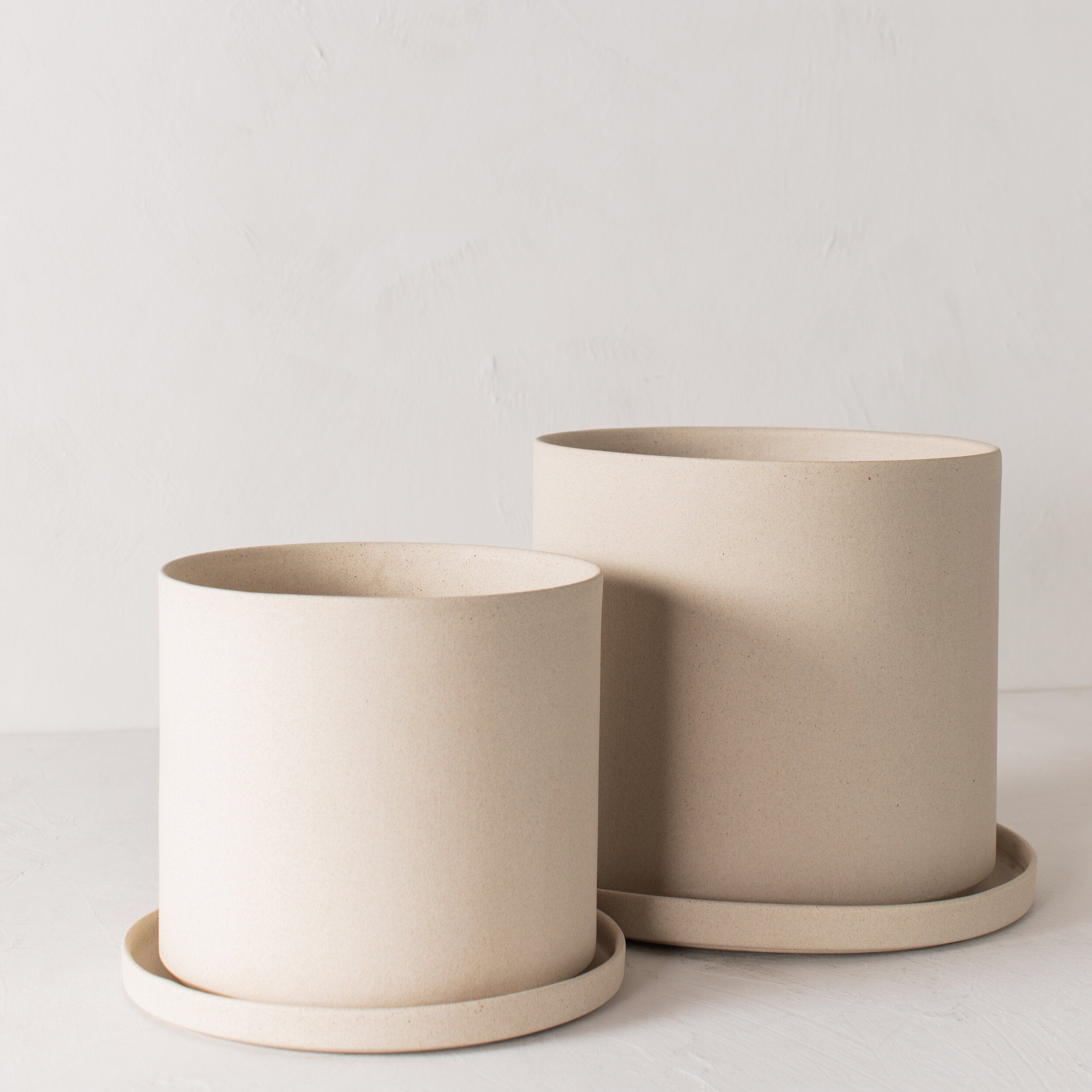 Two stoneware 10 inch ceramic planters with bottom drainage dish. Staged on a white plaster textured tabletop against a plaster textured white wall. Designed and sold by Convivial Production, Kansas City Ceramics. 