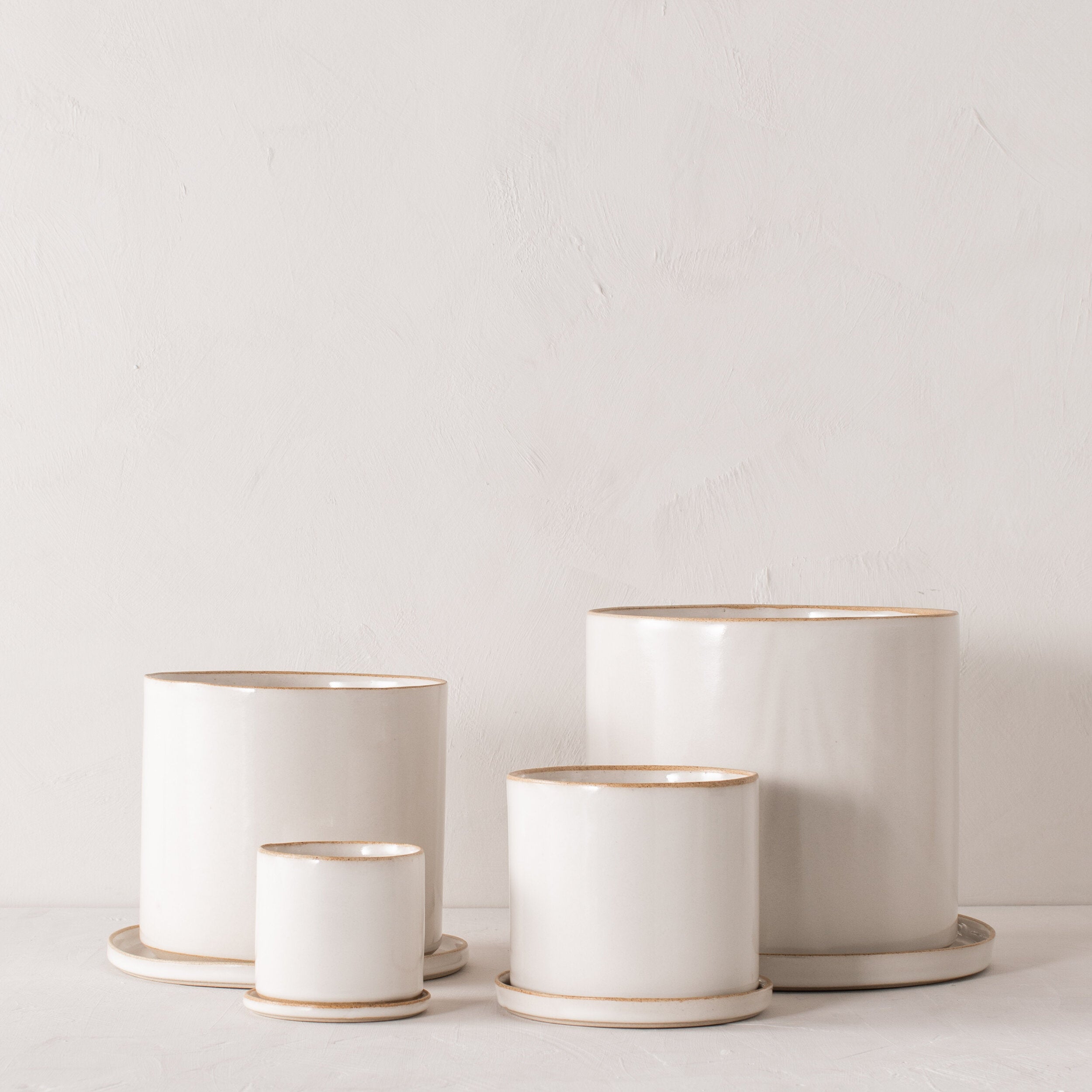 Four white ceramic planters with bottom drainage dishes, 4, 6, 8, and 10 inches. Staged on a white plaster textured tabletop against a plaster textured white wall. Designed and sold by Convivial Production, Kansas City Ceramics.  Edit alt text